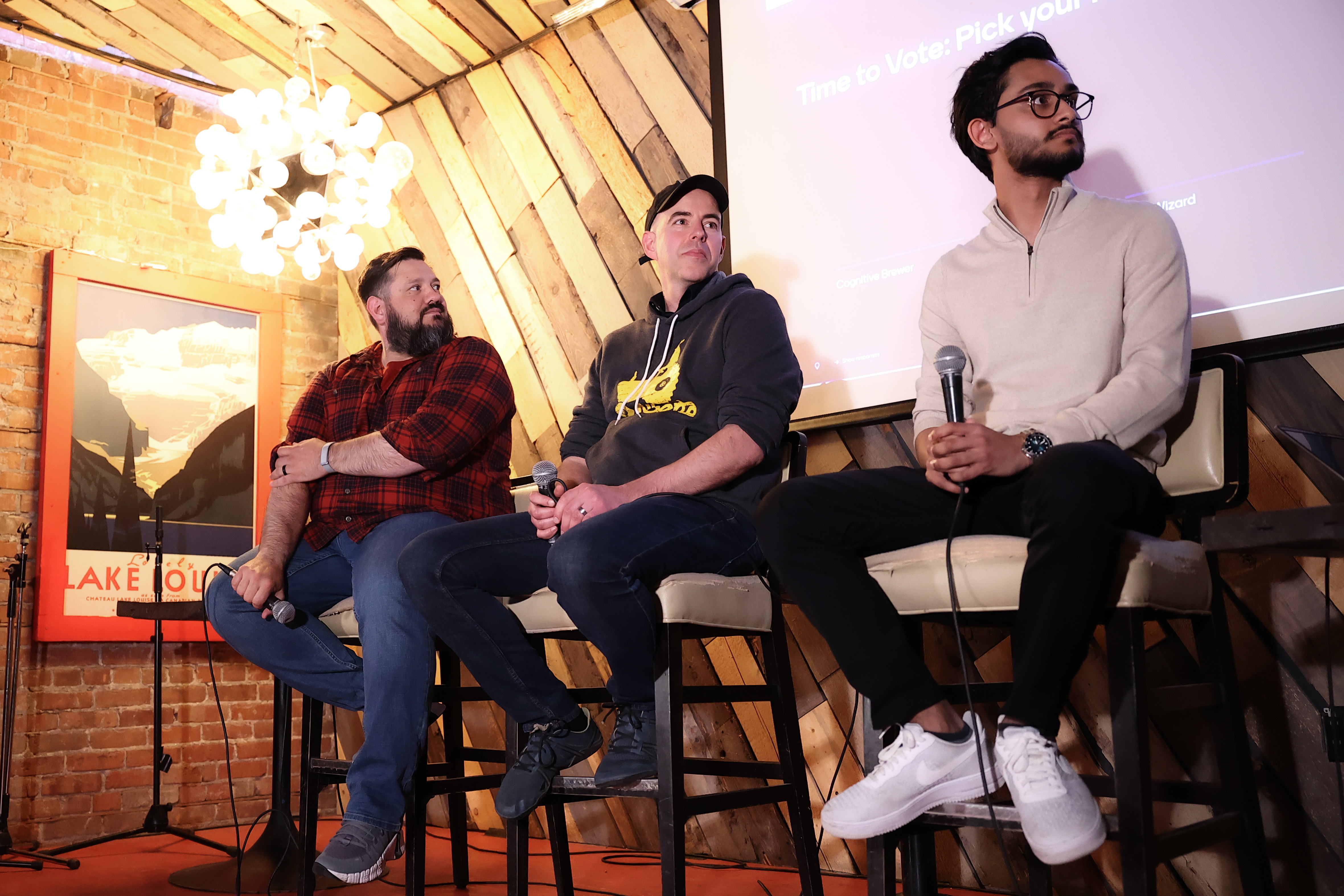 The three competing developers (Rob Edwards, Janak Alford, and Siddhant Gautam) sitting on high barstools on stage, holding microphones, and looking engaged during the Q&A and voting session